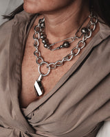 A close-up of a person wearing a layered silver necklace set, featuring a chunky chain with a polished geometric pendant, a beaded chain, and a safety-pin style link, styled with a taupe blouse.