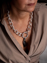 A close-up of a woman wearing a chunky silver chain necklace with a dark faceted pendant, paired with a taupe-brown wrap blouse.