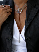 A close-up of a person wearing a silver lariat necklace with a double-ring top and a textured drop pendant, styled with a white top and a dark gray blazer.