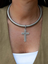 A close-up of a silver snake chain choker necklace with a beaded cross pendant, worn by a model in a khaki blazer over a white top.