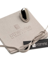 A polished metallic oval-shaped personal sharpener resting on a beige suede-like branded pouch with a ribbon and a black Otherwise tag, photographed on a clean white background.