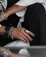 A woman wearing layered silver statement jewelry, including a large pendant necklace, bracelets, and rings, styled with a black vest and white blouse while sitting on a white sofa.