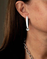 A close-up of a model's ear wearing a polished silver drop earring with a teardrop-shaped top and a small circular dangling accent, paired with a silver chain necklace.