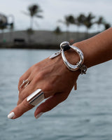 A close-up of a person's hand and wrist wearing a chunky silver ring bracelet with a large open oval and chain details, along with matching silver rings, set against a blurred background of water and palm trees.