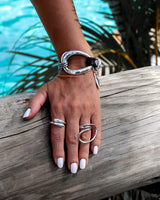 A hand with white nail polish resting on a weathered wooden log, wearing a silver wrap bracelet and multiple silver rings, with turquoise water and palm leaves blurred in the background.