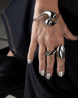 A close-up of a hand wearing multiple sculptural silver rings and a silver wrap bracelet, resting against dark navy fabric.
