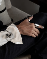 A close-up of a person's hand resting on dark trousers, wearing multiple polished silver rings and a silver ring on the wrist, with a white shirt cuff visible.