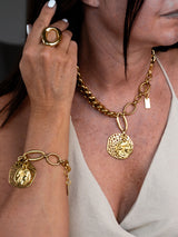 A close-up lifestyle shot of a model wearing a gold chain necklace with a textured circular pendant, a matching gold bracelet with a medallion charm, and a gold ring on her finger, paired with a light beige top.