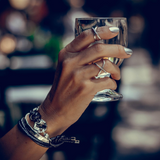 A person's hand holding a glass, wearing a silver edgy punk ring with a 'X' shape on the finger.
