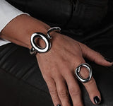 Sculptural wide cuff bracelet with exaggerated fluid loops, featuring a high-shine metallic finish, worn on a model's wrist.
