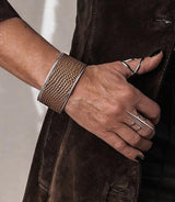 Wide brass bangle featuring beige animal print leather inlaid across a textured band, worn on a wrist with silver rings.