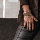 Wide brass bangle featuring beige animal print leather, worn on the wrist, paired with silver rings and sleek black pants.