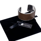 Wide brass bangle featuring a beige-toned animal print leather inlay on a textured band, presented on a black suede pouch.