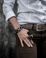 Edgy leather statement bracelet with silver-plated metal design, worn with a white shirt and brown pants.