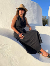 Woman in a black dress and hat sitting on a white wall with a blue sky background