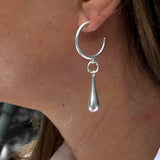 Chunky sterling silver hoop earrings featuring a bold teardrop charm, worn on a model's ear.