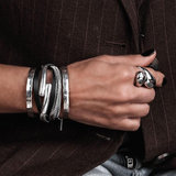 Hand wearing multiple silver bracelets and a ring, with a brown pinstripe coat in the background.