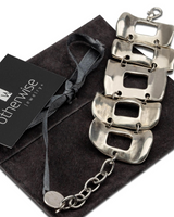 Wide silver-plated zamak arm cuff featuring five irregular metal pieces, adjustable with an extension chain, presented on a suede pouch.