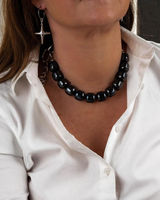 A close-up of a woman wearing a chunky black beaded collar necklace and a silver dangling star earring, wearing a white button-down shirt.