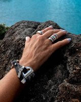 Bold woven leather cuff bracelet with metal snake chains, worn on a hand, showcasing layered, edgy style against a rocky background.