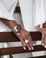 Woven leather and metallic snake chains create a bold cuff bracelet, worn on a wrist with a ring, against a white outfit.