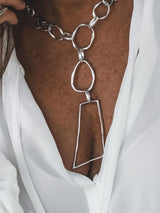 Chunky asymmetrical silver plated necklace with large irregular pendant and carabiner clasp, styled over a white blouse.
