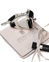 Bold stirrup leather and metal bracelet featuring black leather cords and a shiny silver stirrup motif, resting on a suede pouch.
