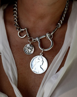 A close-up of a silver chain necklace worn by a person. The necklace features a large toggle clasp, a smaller ring clasp, and two coin-style charms, including a prominent silver dollar charm.