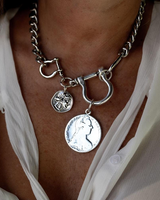A close-up of a silver chain necklace with a large toggle clasp and two coin-style charms, worn on a person's neck over a white blouse.