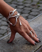 Chunky silver metal cuff bracelet with sculptural link shapes, worn on a wrist with a matching ring, displayed against a stone surface.