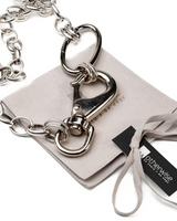 A silver-tone metal chain accessory with a large lobster clasp and D-ring, laid on top of a light beige velvet pouch and ribbon. A black tag reads 'otherwise'.