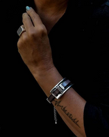 A close-up of a person's wrist wearing a silver-toned metal cuff bracelet with a dark strap, paired with a silver ring, against a dark black shirt.