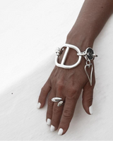 Chunky silver heart bracelet with a large D-shaped design, paired with a sleek ring, worn on a hand with white nails.