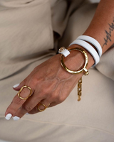 A close-up of a person's hand and wrist wearing multiple jewelry pieces: a large hammered gold ring, a smaller gold ring, a white bangle bracelet, and a gold bangle with a white wrap detail. The person is wearing beige clothing.