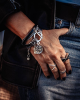A close-up of a person's hand resting on their jeans, showcasing a stack of silver-toned jewelry including a cuff bracelet with a skull charm, a ring, and a geometric band, alongside a black leather jacket.