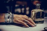 Funky silver-plated metal ring worn among stacked bracelets on a hand with metallic nail polish, resting on a table.