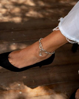 Thick stainless steel chain anklet worn on the ankle, featuring oval links and a charm, paired with a black heel.