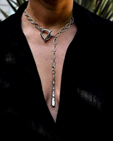 A close-up of a person wearing a silver-toned chain necklace with a toggle clasp and a long vertical pendant, styled with a black open-collar top.