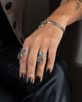 Hand with multiple silver rings and black nail polish wearing a bracelet, sitting on dark fabric.