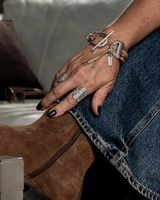 A close-up lifestyle shot of a person's hand resting on their leg, wearing a brown suede ankle boot, blue denim jeans, silver rings, and a silver bracelet with a leather strap and small tag.
