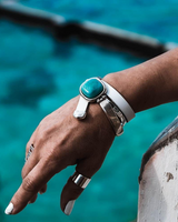 A close-up of a person's hand wearing a wide silver cuff bracelet with a large turquoise stone, along with other silver rings and a silver bangle, set against a blurred turquoise water background.