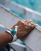 Hand wearing a turquoise stone bracelet and multiple rings on a railing with water in the background