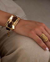 A close-up lifestyle shot of a person's hand resting on their lap, showcasing a chunky gold-plated cuff bracelet with a brown leather strap and a matching gold ring.