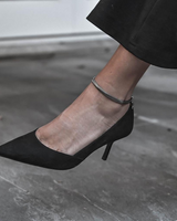 Silver snake chain anklet worn on ankle, paired with a black pointed-toe heel, on a stone surface.