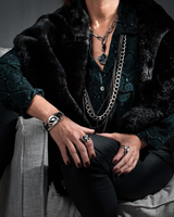 Person wearing a dark coat with fur trim, multiple necklaces, and rings sitting on a checkered chair.