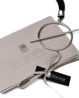 A silver-tone circular ring keychain with a straight bar, presented on top of a light taupe fabric pouch branded 'otherwise / be sharp & be edgy' with a black ribbon tag, on a white background.