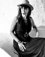 Black and white photo of a woman wearing a wide-brimmed hat and sunglasses, sitting against a light-colored wall.