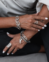 A close-up of a person's hands and arms wearing multiple silver-toned jewelry pieces, including bracelets, rings, and a large silver ring, styled with a light gray top and black pants.