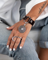 Brown leather cuff bracelet featuring a horse bit buckle and a circular silver pendant with a white opal crystal.