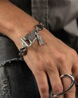 A close-up of a person's hand and wrist wearing a silver-toned chain bracelet with padlock and key charms, along with a large silver ring on the finger, set against gray denim clothing.